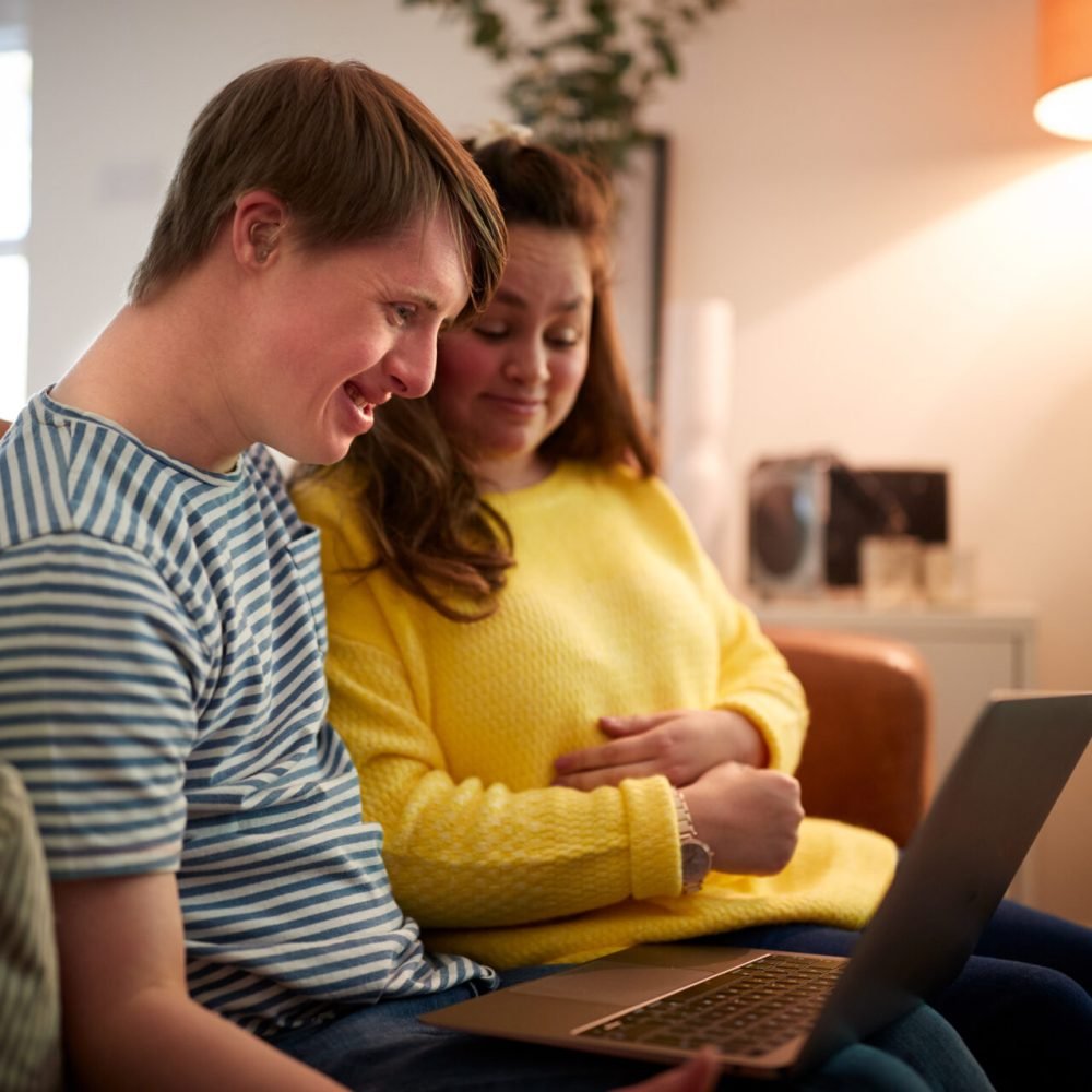 Young Downs Syndrome Couple Sitting On Sofa Using Laptop At Home